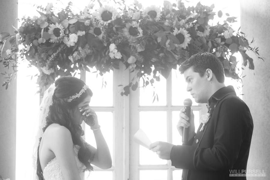 Couple sharing vows at their wedding ceremony; the groom is reading his vows into a mic while the bride wipes a tear from her eye