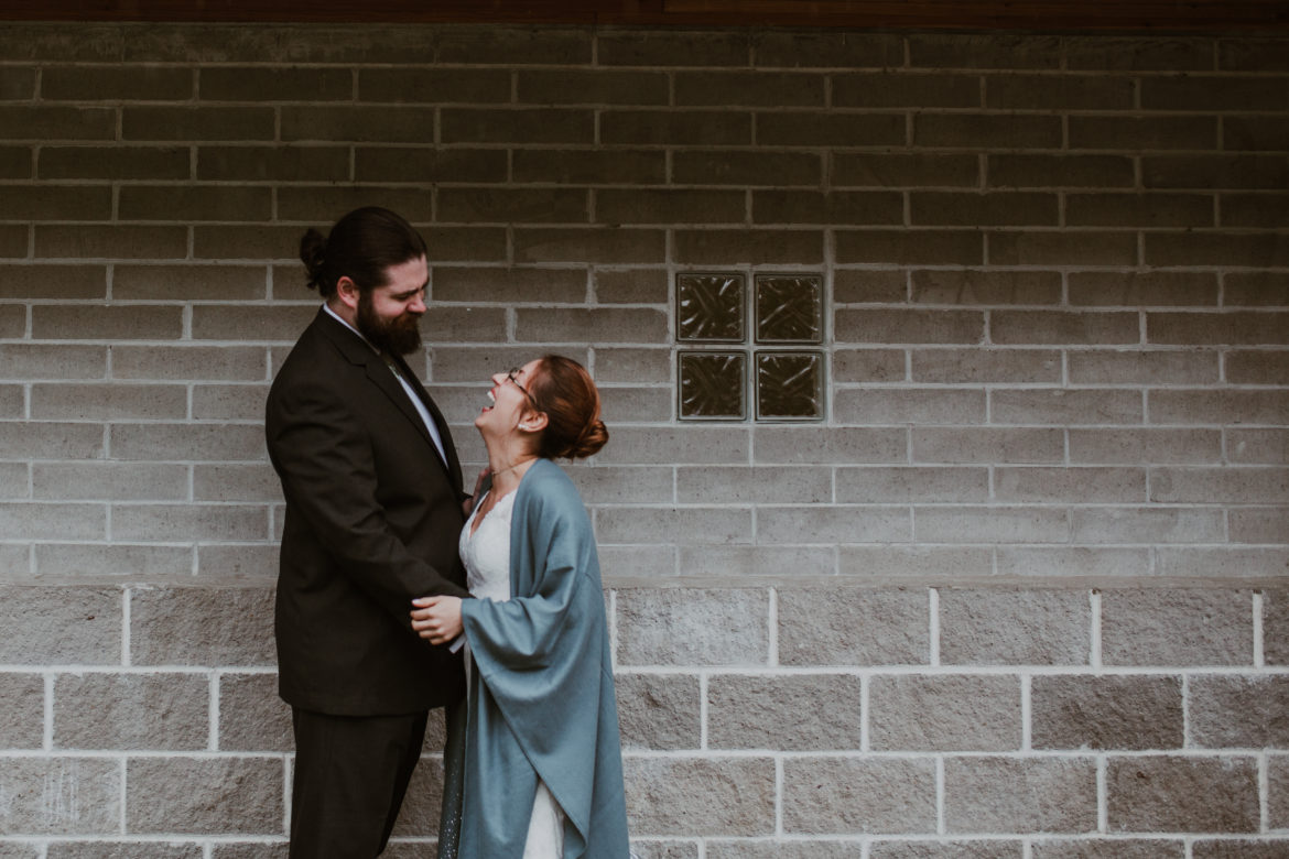 Newlywed couple smile at each other while holding hands in front of a brick wall