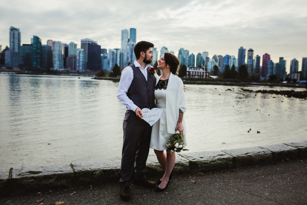 Newlywed couple on the Vancouver seawall with the city skyline behind them holding a heart that says they eloped